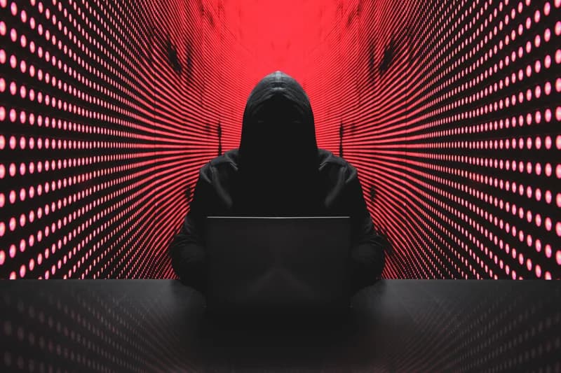The image depicts a person in a hooded sweatshirt sitting in front of a laptop. The person's face is obscured by the hood. The background is a pattern of red lights, creating a tunnel-like effect.