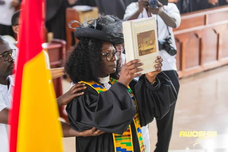 A female Presbyterian minister wearing ceremonial black robes and a kente-patterned stole holds a large Bible with an image of 'The Last Supper' on its cover. She is being assisted by others during the official inauguration ceremony of the Awoshie District within a church setting.