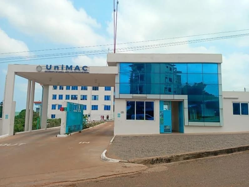 A wide-angle shot of the main entrance to the University of Media, Arts and Communication (UniMAC) campus. The scene features a modern security post with reflective blue glass windows and a gated driveway leading towards a multi-story white building in the background.