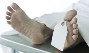 The image depicts the feet of a deceased individual resting on a metallic surface, likely a morgue table. A blank white identification tag is tied to the big toe, and the body is partially covered by a white shroud or sheet.
