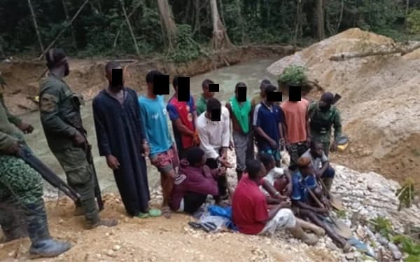 A group of thirteen young men, suspected of illegal mining, are held by armed security personnel in a forested area. The scene features heavily excavated earth and a muddy water body, typical of a 'galamsey' site in Ghana. The suspects' faces are blacked out, and they are being guarded by officers in camouflage uniforms.