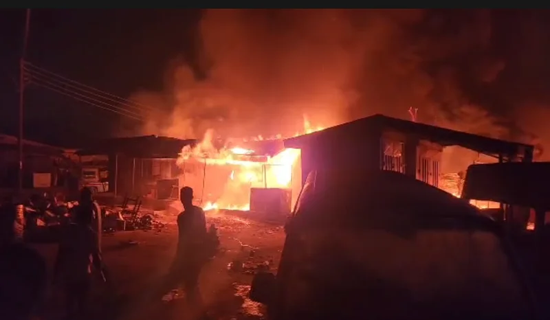 A massive fire is shown engulfing several structures and shops at night, with thick smoke rising into the air. Silhouetted figures of people are visible in the foreground observing the inferno at what is reported as Sofoline Magazine.