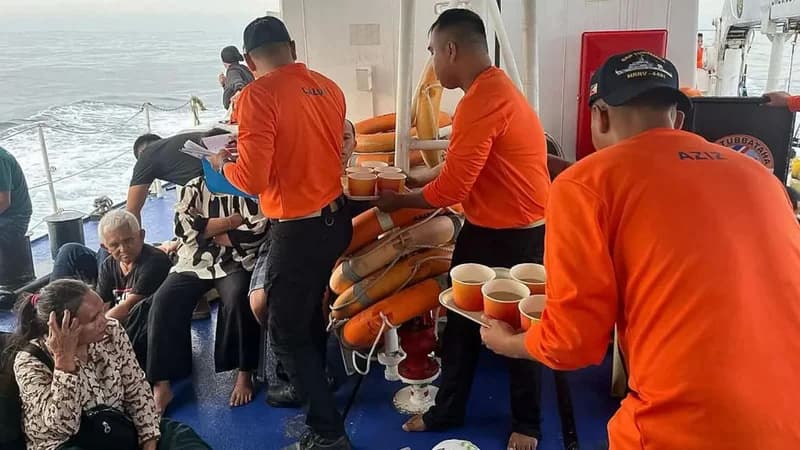 Rescue personnel in bright orange uniforms are seen providing assistance to survivors on the deck of a ship after a ferry disaster. The image shows workers distributing drinks in cups to distressed individuals sitting on the floor near a stack of life rings.