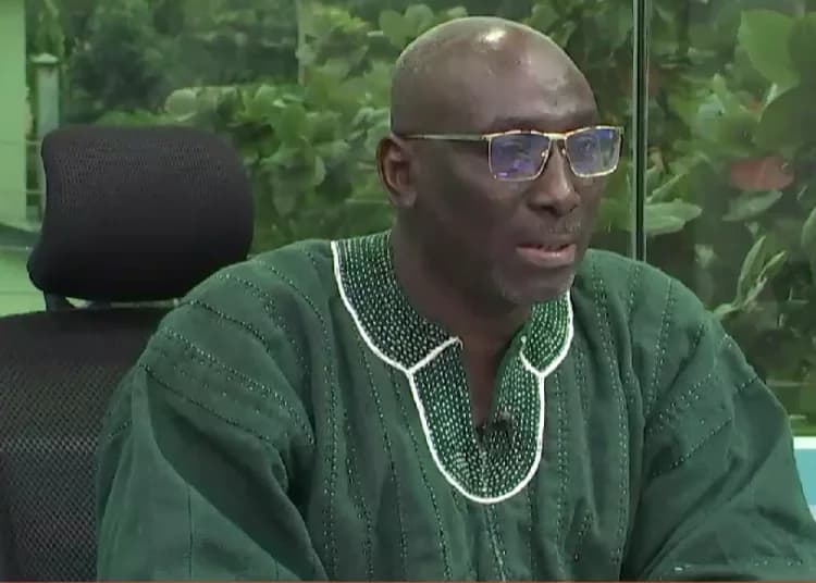 A man wearing a traditional green Ghanaian smock (fugu) and gold-rimmed glasses is pictured in a studio or office setting. He is seated in a black chair with a headrest, appearing to be in the middle of a discussion or media interview.