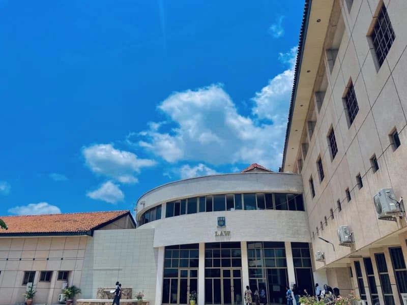 A bright daytime photograph of the University of Ghana School of Law building in Legon, Accra. The facade features a prominent curved section with a large glass entrance, the university crest, and the word 'LAW' displayed clearly above the doors.