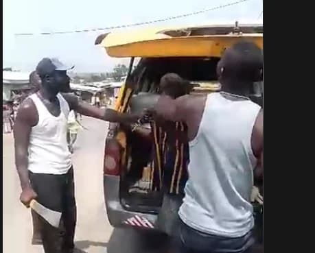 The image shows a tense scene where a man in a white tank top and cap is holding a large machete near a yellow commercial vehicle. Another man is seen assisting someone at the rear of the same vehicle, which has its trunk open. The setting appears to be an outdoor public area, capturing a moment during a reported violent confrontation.