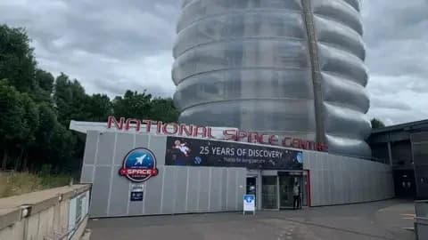 The photograph shows the front of the National Space Centre in Leicester with a banner which reads "25 years of discovery". It is a grey building with a rocket tower.