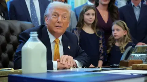 Getty Images Trump, in a blue suit and yellow tie, sits at his desk in the Oval Office. There are people standing around hi, including two young girls to his left. On his desk is a large bottle filled with milk.