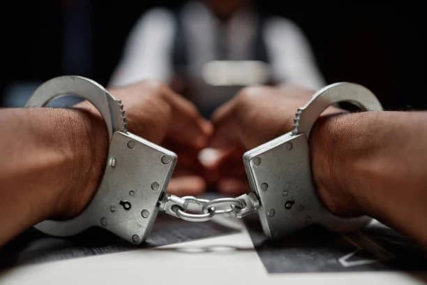A close-up shot of a pair of metal handcuffs locked around the wrists of a person whose hands are resting on a surface. In the background, the blurred figure of another person is visible, suggesting an interrogation or police station setting.