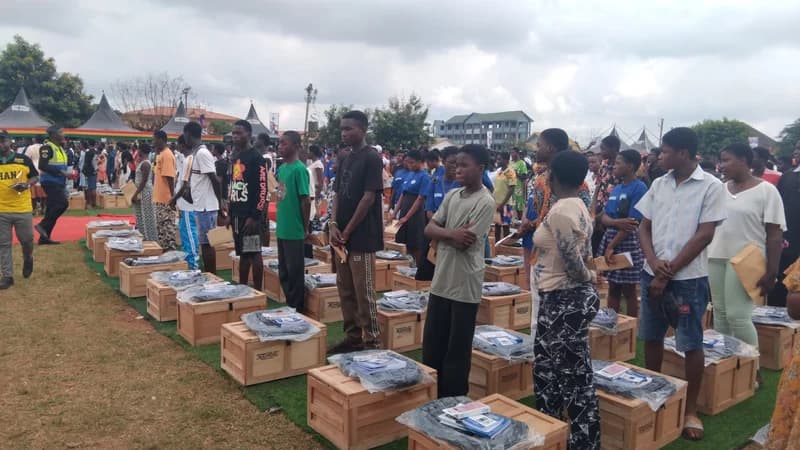 A large group of students is gathered at an outdoor ceremony in Ghana, standing in rows next to wooden trunks and school supplies. Each trunk is topped with a mattress and various educational materials, prepared for distribution to SHS entrants.