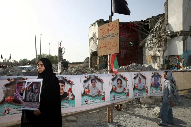 A woman dressed in a black chador holds a framed photograph of two children in front of the remains of a destroyed building. A long banner featuring portraits of several other children serves as a memorial, set against a backdrop of rubble and a heavily damaged structure. The scene depicts the human and physical aftermath of a military strike in a residential or educational area.