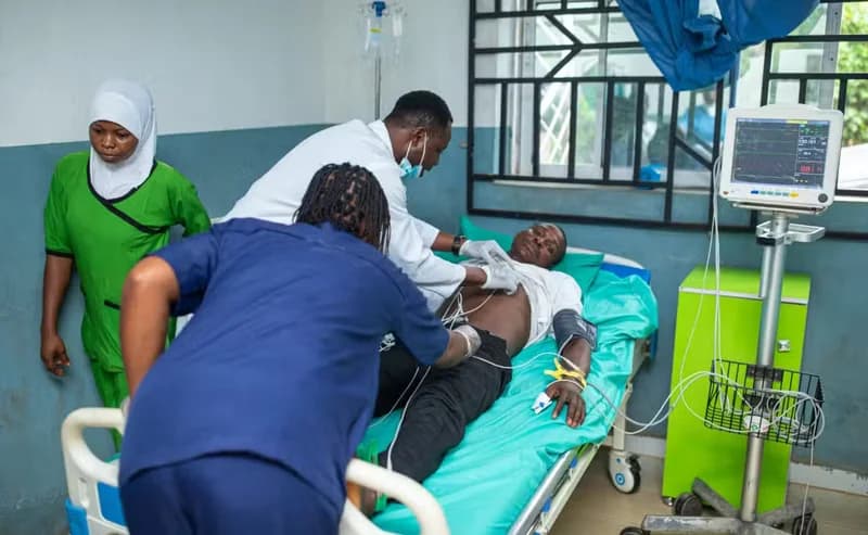 A male patient is being attended to by three healthcare professionals in a hospital setting. The patient lies on a bed connected to medical monitoring equipment while staff in scrubs and white coats perform clinical assessments.