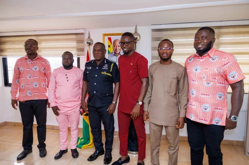 A group of six men, including a senior Ghana Police officer and representatives from the Okada Riders Association of Ghana (ORAG), pose for a photo in an office. A portrait of former President Nana Akufo-Addo and the Ghana flag are visible in the background, indicating a formal state-related meeting. Some men are wearing shirts branded with 'ORAG', identifying their association.