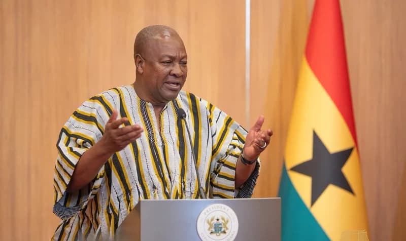 President John Mahama is captured speaking at a podium, gesturing with his hands during an official address. He wears a traditional Ghanaian smock with yellow and black stripes, positioned next to the national flag of Ghana against a wood-paneled background.