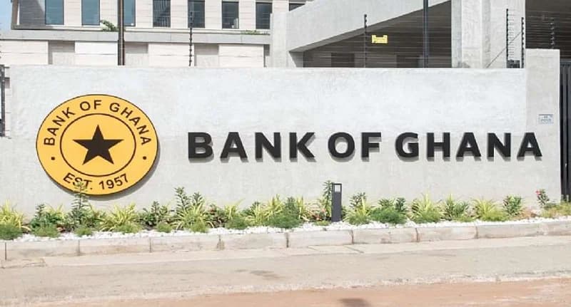 The image shows the exterior wall of the Bank of Ghana, featuring the official seal and the institution's name in bold, black block letters. The gold seal includes a central black star and the text 'EST. 1957'. Small green plants and white stones are neatly landscaped at the base of the light-grey textured wall.