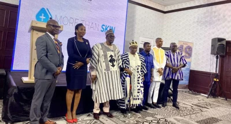 Speaker of Parliament, Alban Bagbin, stands with a group of officials and dignitaries on a stage during a health ceremony. Behind them is a large digital screen with the text 'MORE THAN SKIN' alongside a medical cross logo.