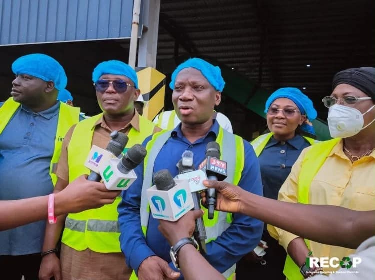 A group of officials wearing neon yellow safety vests and blue hairnets are being interviewed by journalists at what appears to be an industrial facility. The central figure is speaking into multiple microphones from major Ghanaian media outlets like Joy News and Channel One TV during a site inspection.