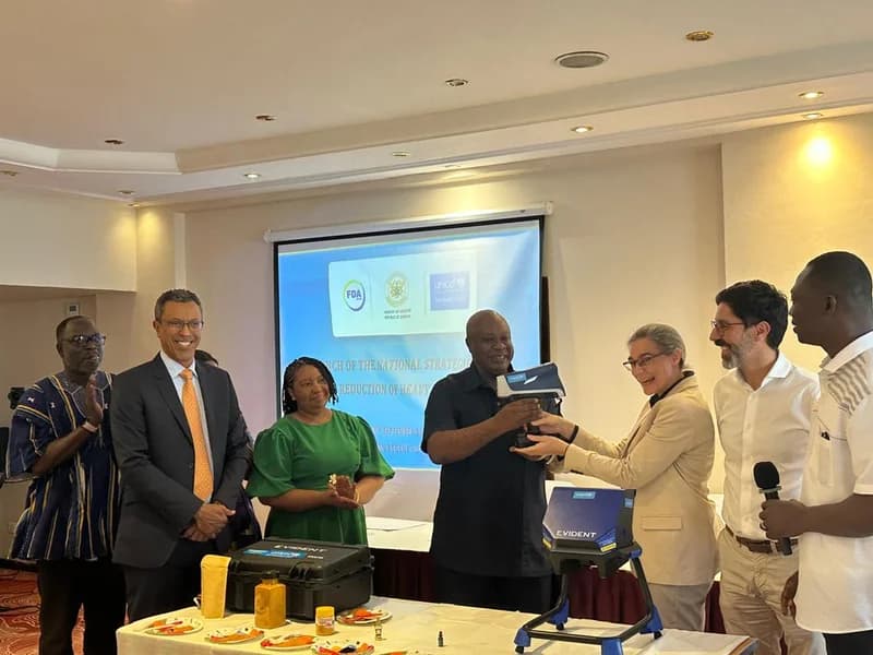 Officials from Ghana's Food and Drugs Authority (FDA), the Ministry of Health, and UNICEF participate in the launch of a national strategic plan to reduce heavy metals in food. A man in a black shirt holds a handheld testing device, likely an XRF analyzer, while standing before a projector screen displaying the event's title and partner logos.