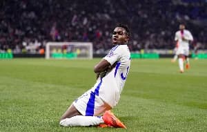 Ghanaian international Ernest Nuamah celebrates on his knees after scoring for Olympique Lyonnais. He is captured in his white and blue club kit during a match at a packed stadium.