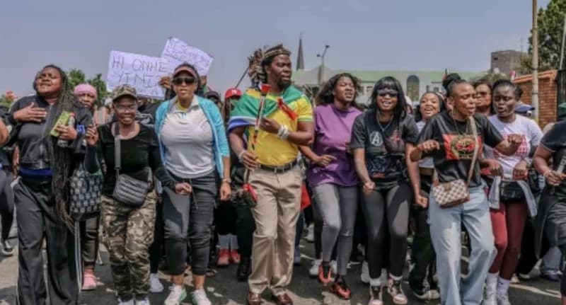 A large group of protesters, primarily women, are seen marching and chanting on a street in South Africa. A man in the center wears a South African flag-themed garment, while others hold placards with handwritten messages demanding an end to violence against women.