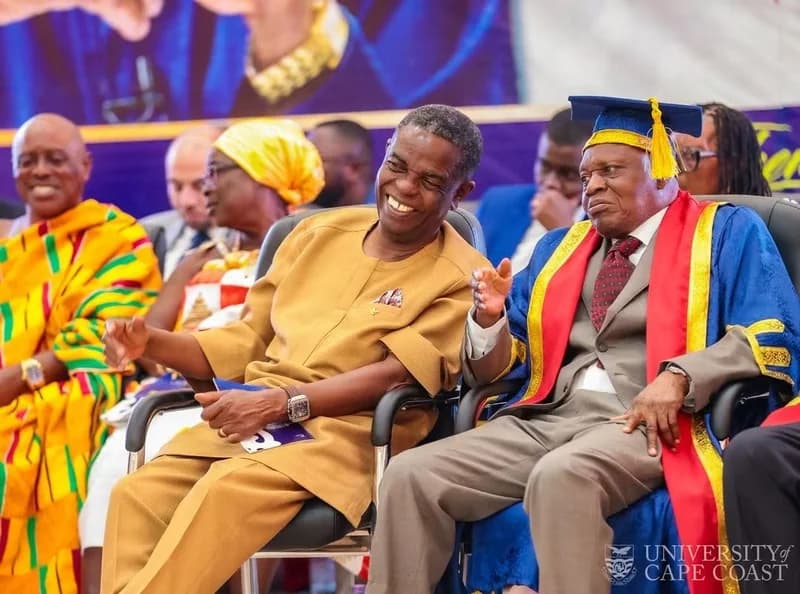 Veteran Ghanaian journalist Kwesi Pratt Jr. is seen laughing while seated next to a dignitary in academic regalia at a University of Cape Coast function. The image captures several prominent figures, including a man in traditional Kente cloth, attending what appears to be a formal ceremony.