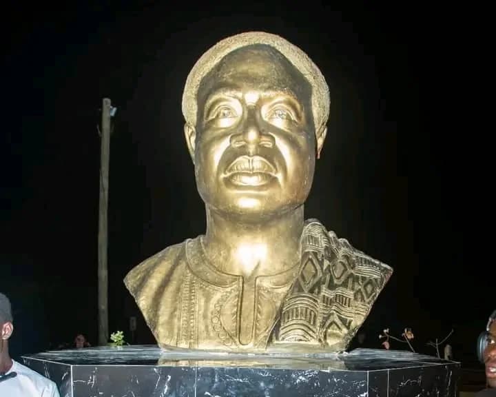 A large gold-colored bust of Dr. Kwame Nkrumah, Ghana's first president, is mounted on a dark, octagonal pedestal. The statue is depicted wearing traditional attire and is illuminated against a dark night background, likely captured during the unveiling event.
