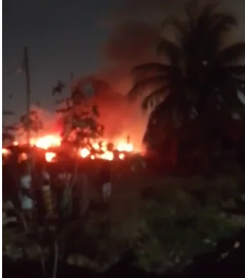A large fire blazes at night, illuminating the surrounding area with a bright orange glow and thick smoke. Silhouettes of onlookers and a palm tree are visible in the foreground against the intense light of the fire.