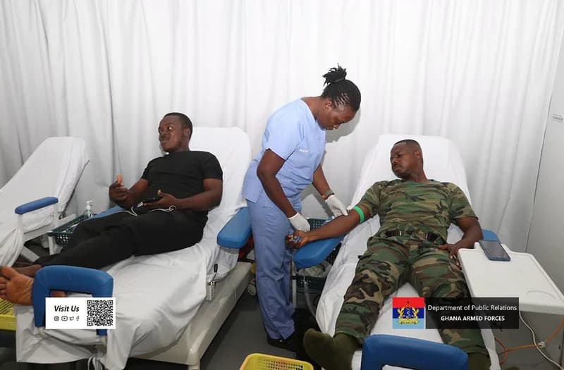 A nurse in blue scrubs attends to a Ghana Armed Forces officer in camouflage uniform during a blood donation drive. Another donor in a black shirt lies on an adjacent bed in a clean, white-curtained medical facility.