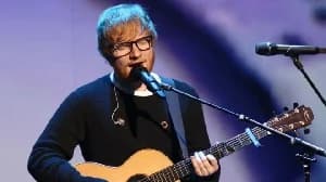 Singer-songwriter Ed Sheeran is captured performing live on stage, playing an acoustic guitar while singing into a microphone. He is wearing his signature black-rimmed glasses and a dark sweater against a softly lit blue and purple background.