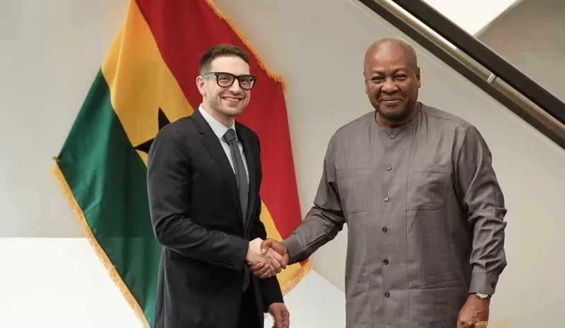 Ghanaian President John Dramani Mahama is pictured shaking hands with Alex Soros, Chairman of the Open Society Foundations. Both men are smiling in front of a prominently displayed Ghana national flag, signifying an official diplomatic or high-level meeting.