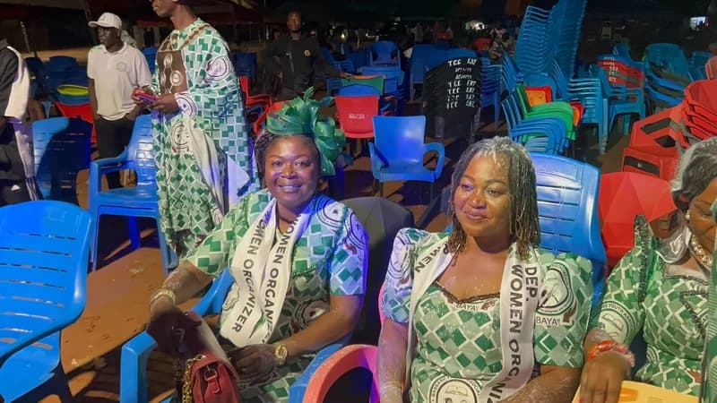 Several women in matching green and white patterned traditional outfits are pictured celebrating at an outdoor event at night. They wear sashes designating them as 'WOMEN ORGANIZER' and 'DEP. WOMEN ORGANIZER' for the Bassari Youth Association (BAYA), with white powder on their clothes and hair signaling a victory celebration.