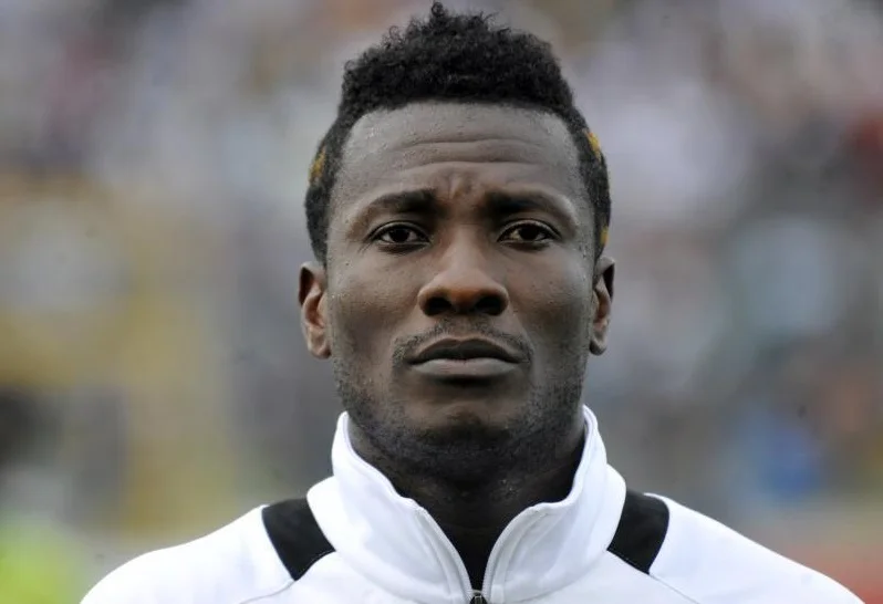 A close-up headshot of Ghanaian football legend Asamoah Gyan. He is wearing a white and black track jacket, and the background shows a blurred stadium crowd, capturing a focused and serious expression.