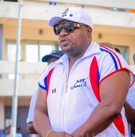 Henry Nana Boakye, popularly known as Nana B, the National Organizer of the New Patriotic Party (NPP), is pictured wearing branded party attire. He is seen outdoors wearing a white sleeveless NPP jacket, a matching cap, and sunglasses, likely during a political event.