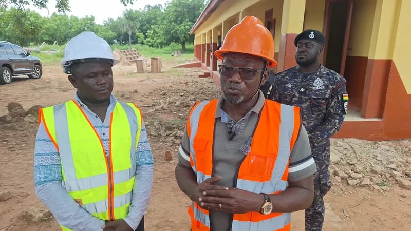 An official, likely the Bono East Minister, wearing an orange hard hat and safety vest, speaks to the camera during an inspection of a school construction site. He is accompanied by another official in a lime green vest and a police officer in camouflage uniform, with a partially completed yellow and brown school building in the background.
