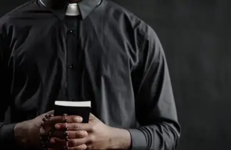 A close-up shot of a person wearing a black clerical shirt with a white Roman collar, holding a small black book and a rosary. The image focuses on the hands and attire of a religious figure against a dark, out-of-focus background.