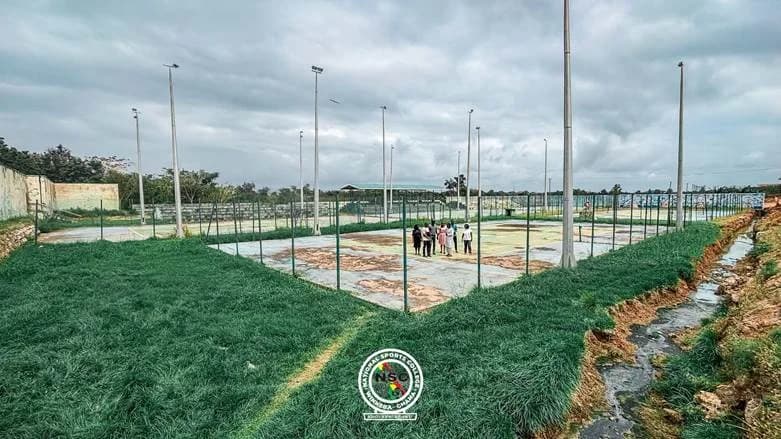 The image shows the outdoor tennis courts at the National Sports College in Winneba, Ghana, which appear to be in a state of disrepair with worn surfaces. A group of individuals is gathered on one of the courts, likely for an inspection or ceremony related to the announced renovation project. The area is surrounded by tall floodlights, fencing, and overgrown grass near a drainage ditch.