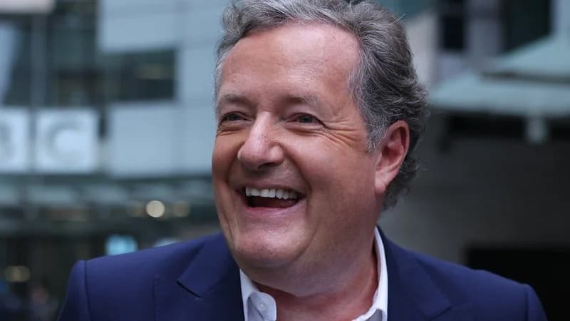 A close-up photograph of British media personality Piers Morgan, shown smiling and laughing. He is wearing a dark blue suit jacket over a white shirt, with a blurred background that appears to be the exterior of a BBC building.