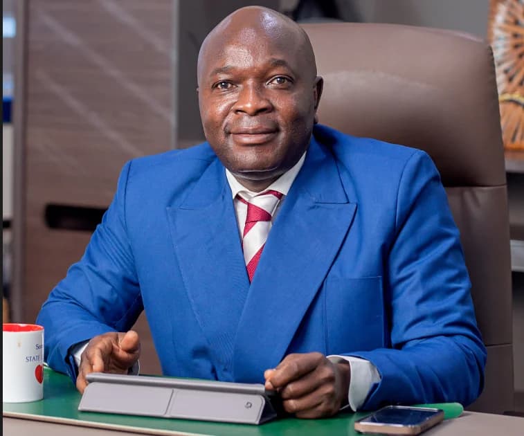A professional portrait of a man in a vibrant blue suit and a red-and-white striped tie, seated at a desk in an office setting. He is positioned behind a tablet and a smartphone, with a branded mug to his left, looking directly at the camera with a composed expression.
