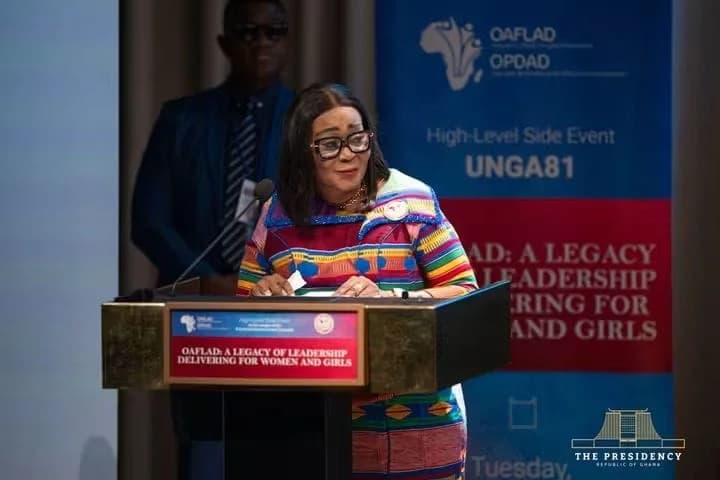 The First Lady of Ghana is pictured delivering a speech at a podium during a high-level side event for the Organization of African First Ladies for Development (OAFLAD) at UNGA81. She is wearing a vibrant, multi-colored Kente-patterned outfit and glasses, while a blurred official stands in the background against an event banner.