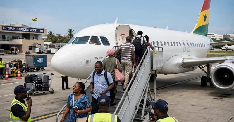 Passengers are seen boarding and disembarking a white aircraft at Kotoka International Airport in Accra. The scene captures the daily activity at Ghana's main aviation hub, with ground crew in high-visibility vests assisting travelers. The aircraft features the Ghanaian flag on its tail, and the terminal building is clearly visible in the background.