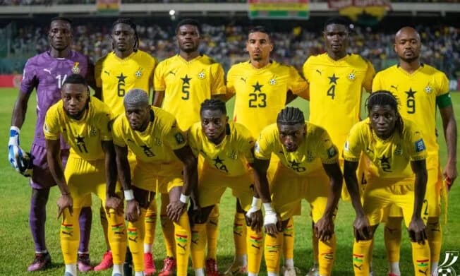 The Ghana national football team, known as the Black Stars, posing for a group lineup photo on a stadium pitch. The players are wearing their yellow Puma jerseys featuring the iconic black star, with the goalkeeper standing on the far left in a purple kit.