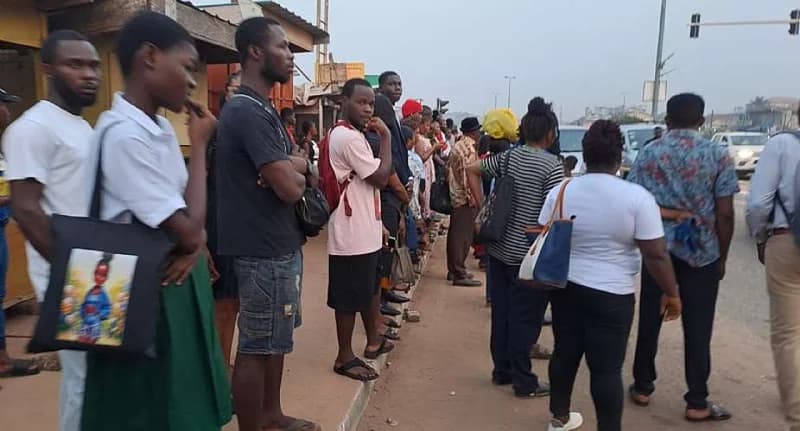 A group of commuters, including students in green uniforms and adults with bags, are seen waiting in a long queue on a sidewalk in Accra. The scene depicts people patiently standing by the roadside, likely waiting for commercial transport like trotros or buses near a busy intersection.