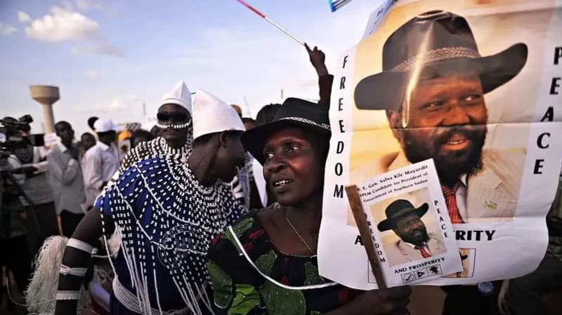 A crowd of supporters at a political event in South Sudan. A woman in the foreground prominently holds a campaign poster featuring President Salva Kiir Mayardit, while others nearby are dressed in traditional attire with elaborate white beadwork.