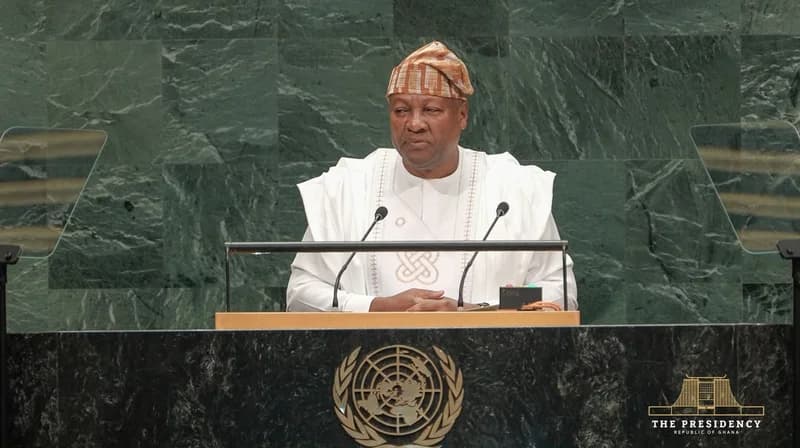 President John Dramani Mahama addresses the United Nations General Assembly, standing at the iconic green marble podium. He is wearing a white traditional outfit and a brown patterned cap, while the United Nations logo is visible on the lectern before him.