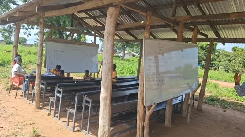 A rural classroom setting in Ghana where students are learning under a makeshift pavilion constructed with wooden pillars and a corrugated metal roof. Two whiteboards are visible, one displaying a mathematics lesson with tally marks and the other an English grammar lesson about nouns, illustrating the infrastructure challenges in the educational sector.