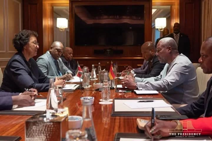 President John Mahama is seen engaging in a formal discussion with a delegation around a polished wooden conference table. The setting features small national flags, including the flag of Ghana, and official stationery, indicating a high-level government or diplomatic meeting.