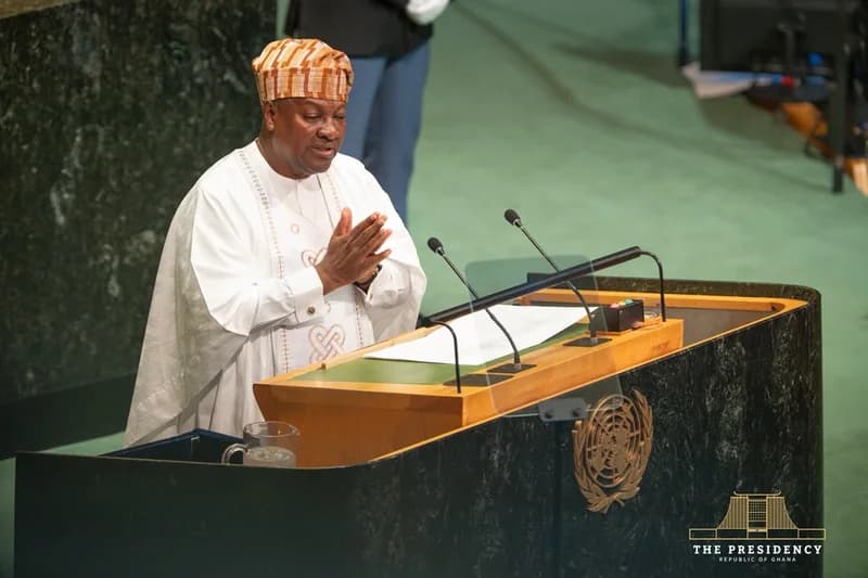 President John Dramani Mahama of Ghana is captured delivering a speech at the United Nations General Assembly. He is dressed in traditional white attire and a patterned cap, standing behind the iconic marble podium which features the UN emblem and a watermark from the Presidency of Ghana.