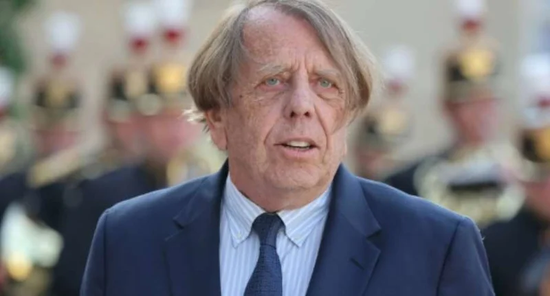 A close-up portrait of veteran football coach Claude Le Roy wearing a navy blue suit and a light-striped shirt. He has a serious expression, and in the blurred background, several individuals in ceremonial uniforms or hats are visible. This photo likely captures the coach during a formal or state event.