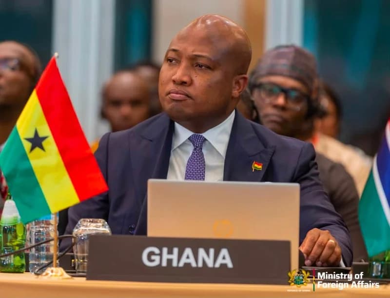 Ghanaian Member of Parliament Samuel Okudzeto Ablakwa is seated at an international summit behind a placard that reads GHANA. He is wearing a sharp dark suit and tie, with a Ghanaian flag and a laptop positioned on the table in front of him.