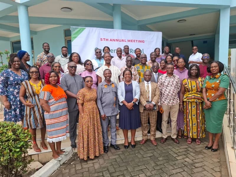 A large group of participants, consisting of men and women in diverse attire, poses for a commemorative photo during the 5th Annual Meeting on Gender and Rural Advisory Services. The group is positioned on a staircase outside a light blue building, standing in front of a white event banner.